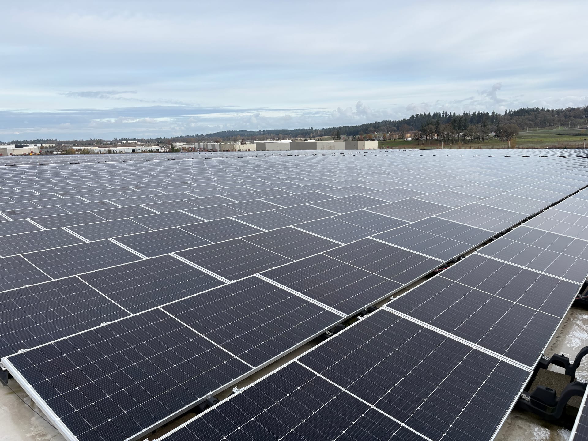 A commercial rooftop solar array stretching to the horizon, warehouses and forested hills beyond