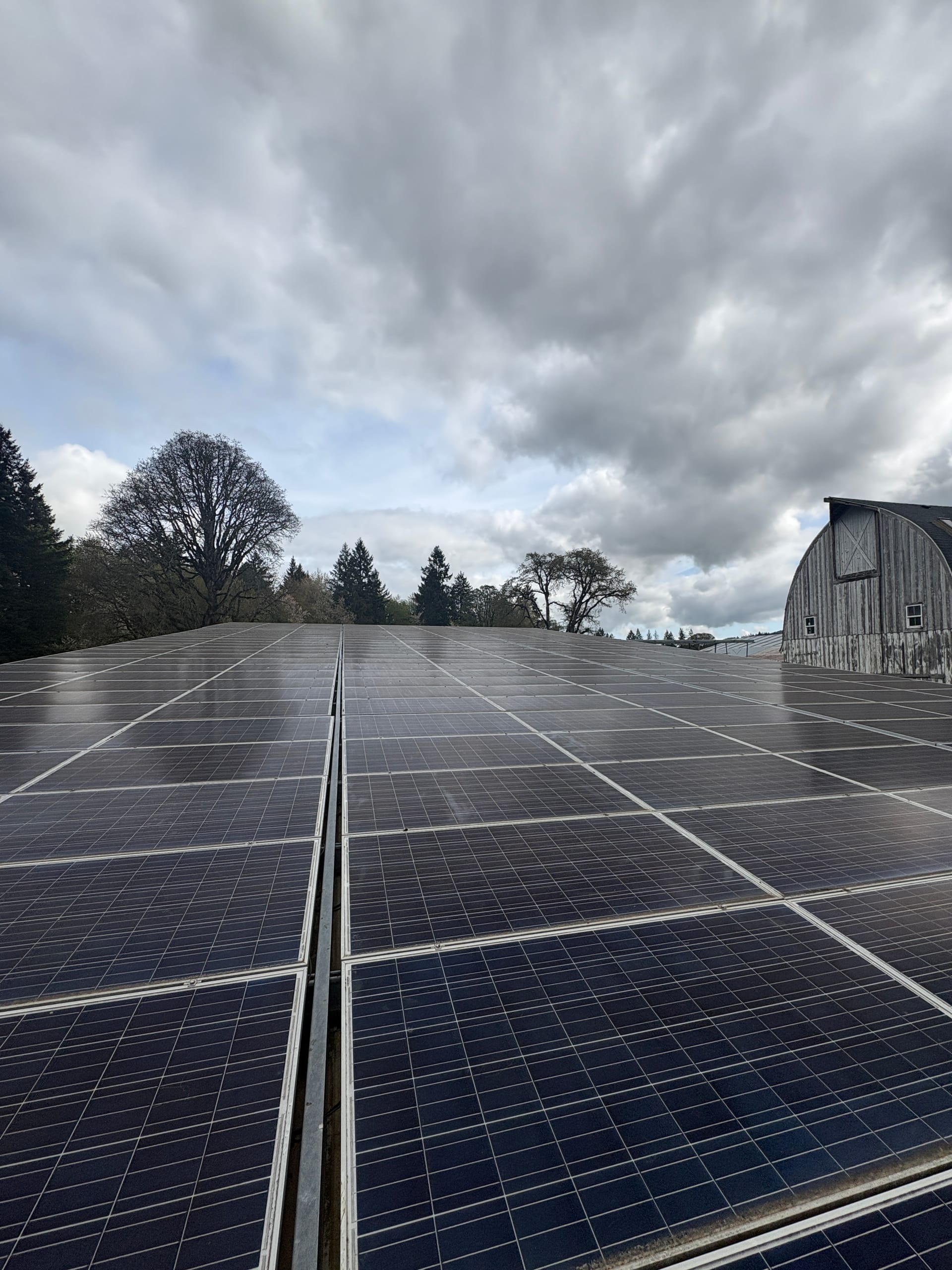 Long rows of a ground-mount solar array beside a weathered barn under breaking storm clouds
