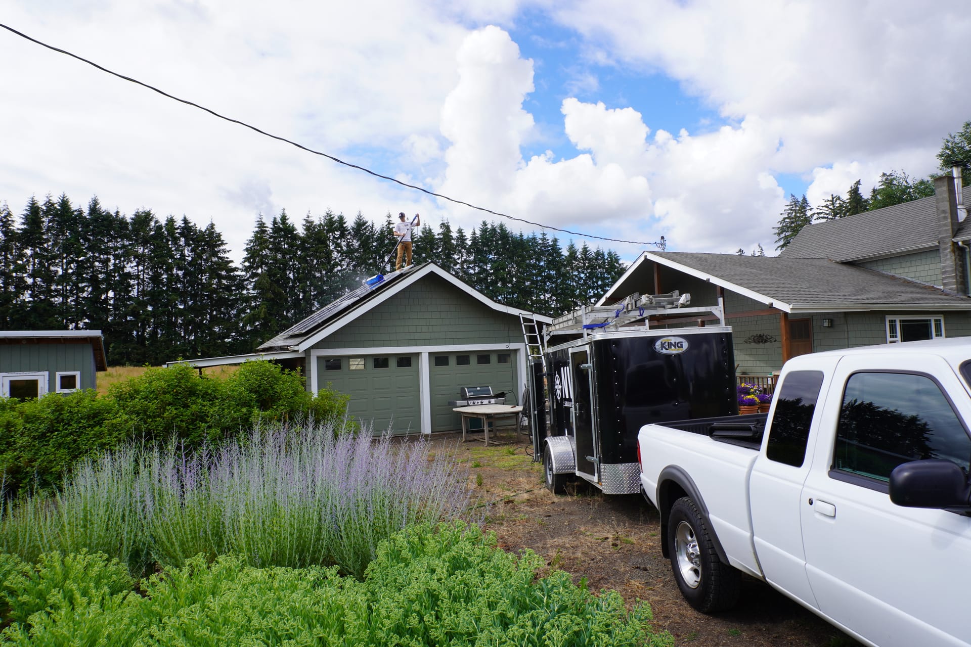 A technician on a garage roof ridge holding a water-fed pole, the brush resting on the panels below