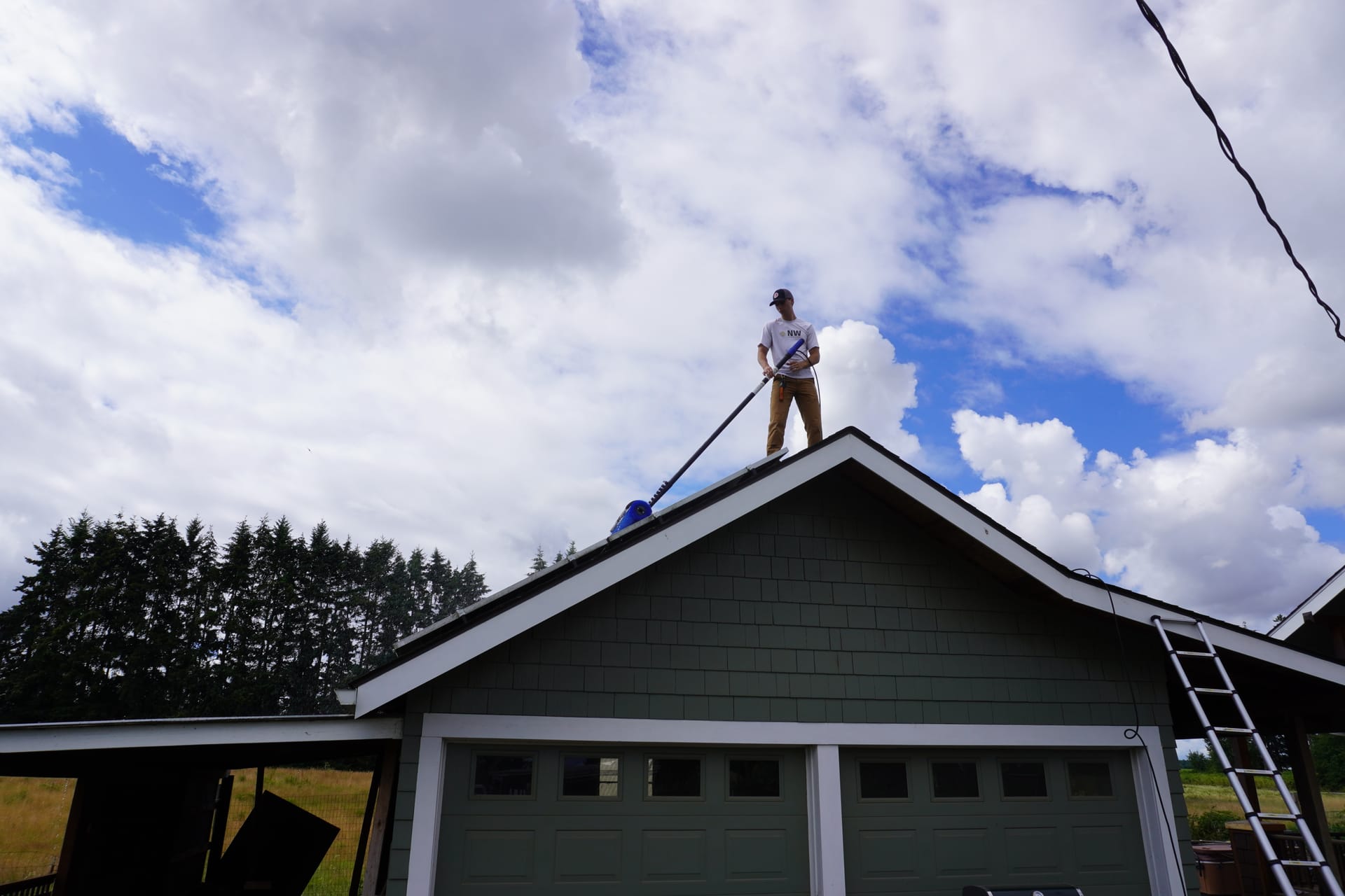 A Northwest Solar Clean technician on a garage roof peak, guiding a water-fed brush across the panels below